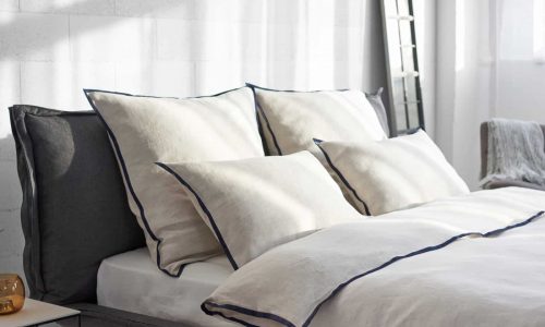 A modern, sun-filled bedroom with a neatly made bed featuring Hirundo linen bedding by Geniksa in cream and navy blue, four large pillows, a duvet and a mirror on the wall.
