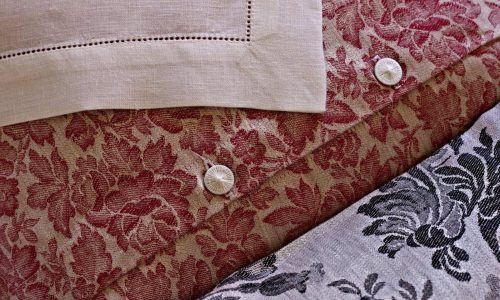 Eine Nahaufnahme von Chrysantheme Rot Leinen Damast von Hoffmann zeigt rote und graue Blumenmuster, zwei weiße Knöpfe und ein Stück beiges Leinen mit Ziernähten in der linken oberen Ecke.