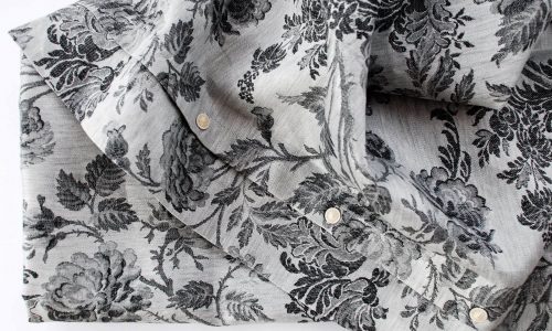 Ein hellgraues, gefaltetes Hemd aus Chrysantheme Anthrazit Leinen Damast von Hoffmann, das ein strukturiertes schwarzes Blumenmuster aus Blättern und Rosen zeigt und mit weißen Knöpfen versehen ist.