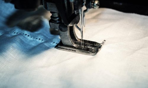 Nahaufnahme einer Nähmaschinennadel, die einen weißen Stoff vernäht, mit detaillierten Fäden und Stoffstruktur beim Niederdrücken des Fußes.