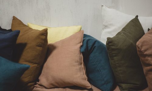Ein Sofa ist mit farbenfrohen Kissen in Blau, Braun, Gelb, Grün, Rosa und Weiß geschmückt, darunter das Hudson Dijon Leinen Coverlet und Kissen von Libeco, die alle vor einer hellen, strukturierten Wand stehen.