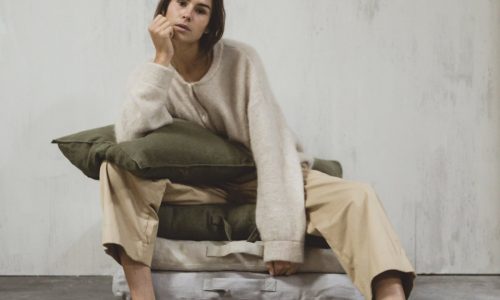 Eine Person, die einen beigen Pullover und eine khakifarbene Hose trägt, sitzt auf gestapelten Hudson Flax Leinen Coverlet und Kissen von Libeco an einer strukturierten Wand, stützt ihr Kinn auf eine Hand und blickt nachdenklich in die Kamera.