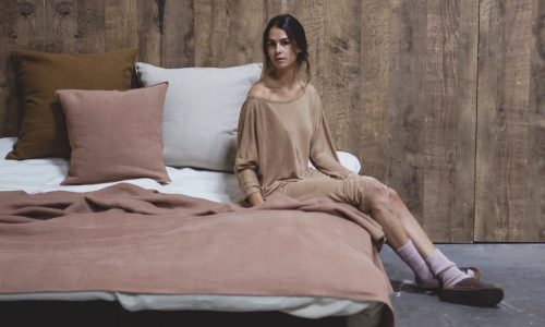 Eine Frau in einem lockeren, beigen Outfit sitzt auf dem Bett mit Hudson Fog Leinen Coverlet und Kissen von Libeco, lehnt sich nach vorne gegen eine rustikale Holzwand und blickt zur Seite.