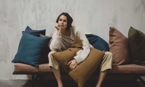 Eine Person sitzt nachdenklich auf einer gepolsterten Bank, die sich an ein braunes Kissen schmiegt, umgeben von großen blauen, braunen und grünen Kissen, vor einer grau strukturierten Wand.