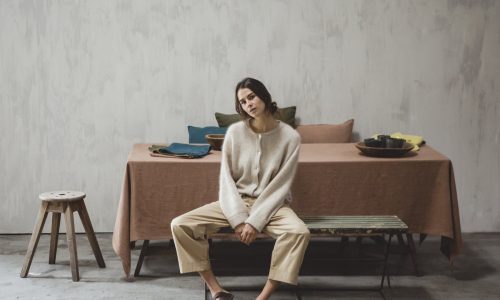 Eine Frau in einem gemütlichen Pullover und einer beigen Hose sitzt an einem mit Hudson Optic White - reines Leinen für den Tisch von Libeco geschmückten Esstisch in einem minimalistischen, neutral getönten Raum mit Betonböden und einer einfachen Kulisse.