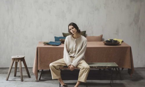 Eine Frau in einem gemütlichen Pullover und einer beigen Hose sitzt an einem mit Hudson Optic White - reines Leinen für den Tisch von Libeco geschmückten Esstisch in einem minimalistischen, neutral getönten Raum mit Betonböden und einer einfachen Kulisse.