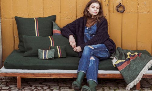 Eine Person in einem dunklen Pullover, einem braunen Oberteil, zerrissenen Jeans und grünen Stiefeln sitzt auf einer grün gepolsterten Bank mit gestreiften Kissen und einer Decke auf einer Kopfsteinpflaster-Terrasse vor einer gelben Holzwand.