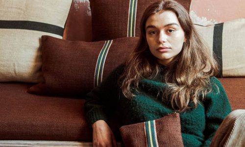 Eine junge Frau mit langen braunen Haaren sitzt auf einem braunen Sofa mit braunen und beigen Kissen hinter ihr. Sie trägt einen dunkelgrünen Pullover, hält eine braune Clutch mit Streifenmuster in der Hand und schaut ruhig in die Kamera.