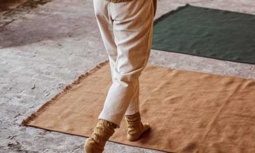 Eine Person, die einen braunen Pullover, eine helle Hose und gelbe Socken trägt, geht auf einem Steinboden, der mit großen rechteckigen Teppichen in Braun-, Hellbraun- und Grüntönen ausgelegt ist. Das Gesicht der Person ist nicht zu erkennen.