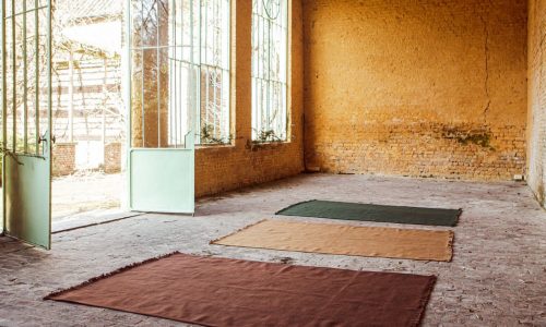 Drei rechteckige Teppiche - braun, hellbraun und grün - liegen auf einem Backsteinboden in einem großen Raum mit gelben Backsteinwänden, großen Fenstern und offenen Türen, die das Sonnenlicht hereinlassen.