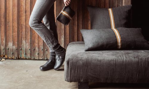 Eine Person in grauen Jeans und schwarzen Schuhen lehnt an einer Holzwand und hält eine kleine dunkle Stofftasche. Zwei dunkle Kissen mit hellbraunen Streifen liegen auf einem passenden niedrigen Sofa in der Nähe.