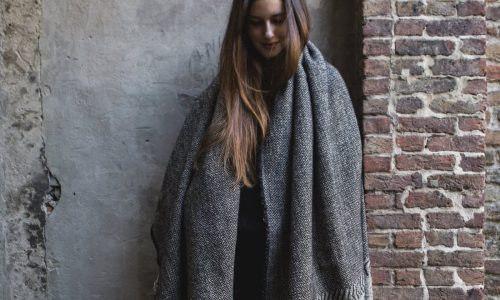 Eine Frau mit langen braunen Haaren steht vor einer strukturierten Wand, eingewickelt in die Decke Jules Black Herringbone Leinen-Wolle-Mix von Libeco, und blickt mit sanftem Blick nach unten.