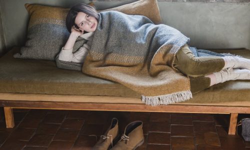 Eine Person entspannt sich auf einem mit der Decke Jules Green Herringbone Leinen-Wolle-Mix von Libeco drapierten Sofa, den Kopf auf einem Kissen, und schaut in die Kamera, während ihre Schuhe ordentlich auf dem gefliesten Boden stehen.