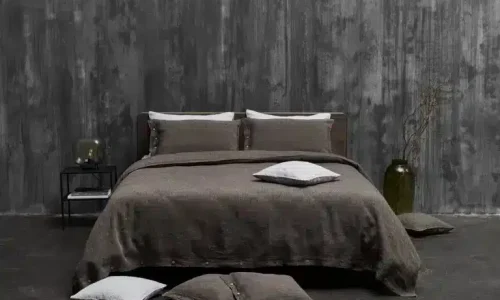 A neatly made bed against a textured grey wall is dressed with luxurious Cygna Classic Linen Sheet by Geniksa, dark brown bedding and matching pillows. On a small side table are a lamp, a glass, and nearby a vase with branches.