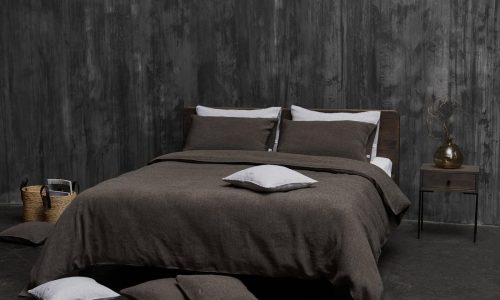 In a modern bedroom, a bed with dark brown Jungle Classic Linen Bedding by Geniksa stands against a textured grey wall. Several pillows lie on the floor, and next to the bed is a small nightstand with a vase.
