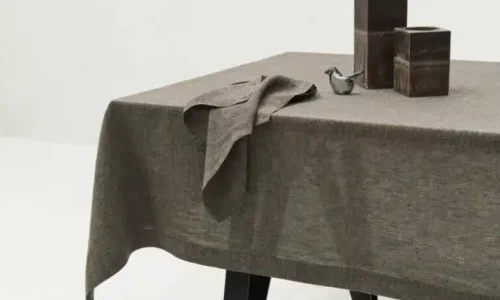 Ein mit einem braunen Tischtuch bedeckter Tisch mit einer gefalteten Serviette, zwei quadratischen dunkelbraunen Vasen und einem kleinen Metallvogel als Ornament auf dem Tisch. Der Tisch steht auf schwarzen Beinen in einem minimalistischen, weißen Raum.