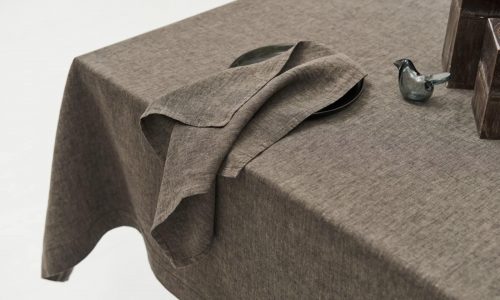 Close-up of a table with the Jungle linen tablecloth by Geniksa, a matching folded napkin on a dark plate, a small glass bird figurine and stacked brown rectangular objects in the corner.