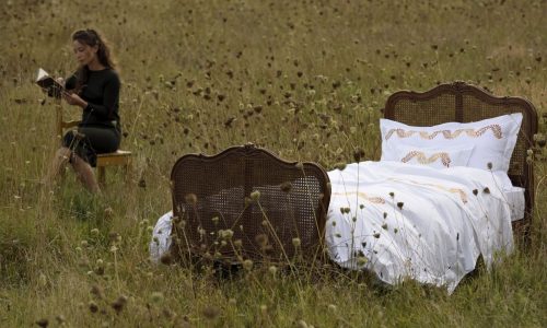 Ein Vintage-Bett, bezogen mit Laurels Percal 400TC Bettwäsche von Peter Reed in Weiß und Gold, steht in einem mit Wildblumen übersäten Feld, während eine Frau in der Nähe auf einem Stuhl sitzt und liest.