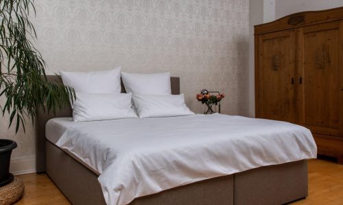 Ein ordentlich gemachtes Bett mit Klassische Weiße Leinen Bettwäsche der Leinenweberei Hoffmann steht in einem hellen Raum mit heller Tapete, Holzboden, großem Holzschrank, blättriger Topfpflanze und einem kleinen Beistelltisch mit einer Blumenvase.