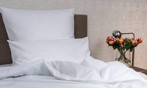 Ein Bett mit weißen Kissen und Weiße Halbleinen Bettwäsche der Leinenweberei Hoffmann steht neben einem hölzernen Nachttisch mit einer Glasvase mit rosa und gelben Rosen vor einer gemusterten cremefarbenen Tapete.