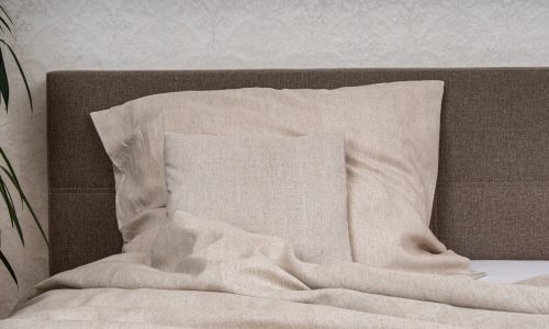 Ein Bett mit beiger Natur Leinen Bettwäsche der Leinenweberei Hoffmann - mit einem großen Kissen, einem quadratischen Kissen und einer Bettdecke - steht vor einem braun gepolsterten Kopfteil und einer weißen Strukturtapete, mit einer Pflanze zur Linken.