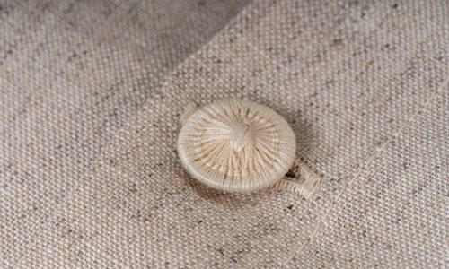 Eine Nahaufnahme zeigt einen beigen stoffbezogenen Knopf, der mit einer feinen Naht auf einen passenden strukturierten Stoff aufgenäht ist - eine ideale Ergänzung zur Natur Leinen Bettwäsche der Leinenweberei Hoffmann.