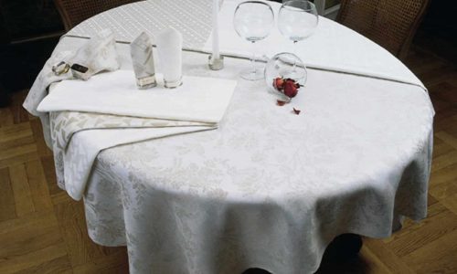 Ein runder Tisch mit der Tischdecke aus feinem weißen Leinen von der Leinenweberei Hoffmann, gedeckt mit zwei leeren Weingläsern, einer brennenden Kerze, gefalteten Servietten, einem Glas mit einer roten Rose und umgeben von zwei Holzstühlen.