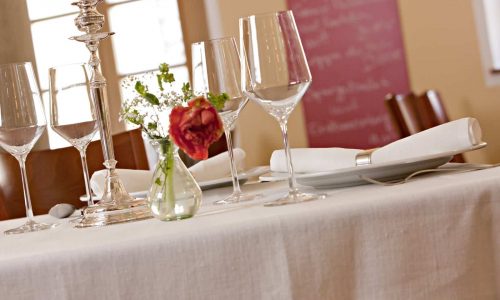 Ein eleganter Restauranttisch ist mit Weingläsern, Tellern, weißen Servietten und Blumen auf einer Tischdecke aus feinem weißen Leinen von der Leinenweberei Hoffmann gedeckt. Im Hintergrund erscheinen Stühle und ein Menübrett sanft verschwommen.