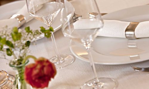 Ein stilvoller Tisch mit weißen Tellern, silbernen Serviettenringen mit weißen Servietten, Weingläsern und einem kleinen Blumenarrangement auf einer Tischdecke aus feinem weißen Leinen von der Leinenweberei Hoffmann.