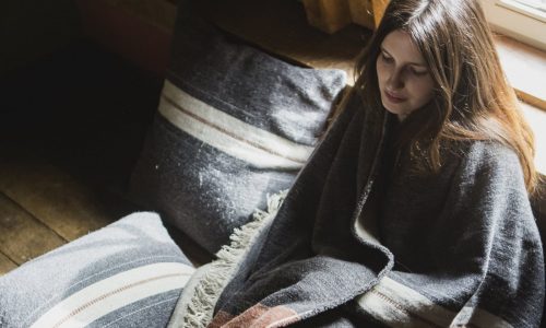 Eine Frau mit langen braunen Haaren sitzt drinnen am Fenster, eingewickelt in die gestreifte Luc Leinen-Wolle-Mix von Libeco-Decke. Neben ihr liegen passende Kissen auf dem Holzboden.