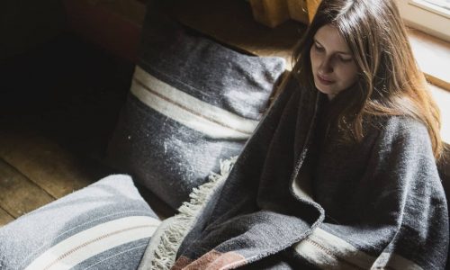 Eine Frau mit langen braunen Haaren sitzt drinnen am Fenster, eingewickelt in die gestreifte Luc Leinen-Wolle-Mix von Libeco-Decke. Neben ihr liegen passende Kissen auf dem Holzboden.