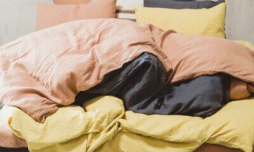 A messy, unmade bed with orange, yellow and gray linens - including Madison Navy colored linens from Libeco - and several pillows against a light-colored wall.
