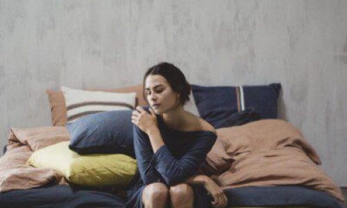 Eine Frau in einem dunklen Kleid sitzt auf der Kante eines Bettes mit farbenfrohen Madison Navy Farbige Leinen Bettwäsche von Libeco und blickt nachdenklich mit einem Arm auf ihrer Schulter auf eine einfarbig strukturierte Wand.