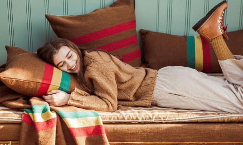 Eine Frau, die einen braunen Pullover, eine beigefarbene Hose und braune Stiefel trägt, liegt auf einer Liege im Freien, schmiegt sich an ein gestreiftes Kissen und lächelt mit geschlossenen Augen. Das Bettzeug und die Kissen sind braun und bunt gestreift.