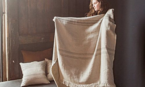 Eine Frau im Haus hält eine große, fransenbesetzte Marrakesch Leinen-Wolle-Mix von Libeco-Decke mit dunklen Streifen hoch. Hinter ihr liegen passende Kissen auf einem Bett und einer Holzwand, während von links Tageslicht einfällt.