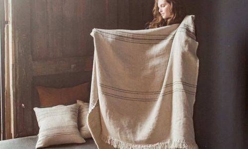 Eine Frau im Haus hält eine große, fransenbesetzte Marrakesch Leinen-Wolle-Mix von Libeco-Decke mit dunklen Streifen hoch. Hinter ihr liegen passende Kissen auf einem Bett und einer Holzwand, während von links Tageslicht einfällt.