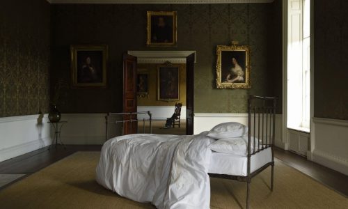 Ein ordentlich gemachtes Bett mit der Matisse Percal 400TC Bettwäsche von Peter Reed steht in einem großen, schwach beleuchteten Raum mit verzierten Wänden und Porträts in Goldrahmen, die ein elegantes, historisches Ambiente schaffen.