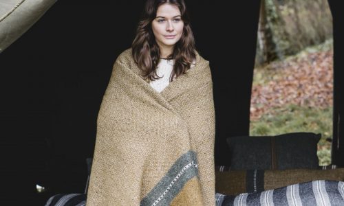 Eine Frau mit gewelltem braunem Haar steht vor einem Zelt, eingewickelt in die Montana Gold Leinen-Wolle-Mix von Libeco Decke, mit Bettzeug hinter ihr und Herbstlaub draußen.