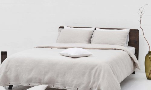 A modern bed with beige linen bedding and pillows stands in front of a white wall. Two pillows lie on the bed, three more are scattered on the white floor, and nearby stands a large green vase with a dry branch.