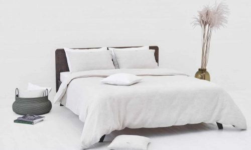 A minimalist bedroom with a neatly made bed in Natural Classic Linen Bedding by Geniksa, two pillows, a white basket with cushions, books on the floor, tall pampas grass in a vase and calming neutral tones.