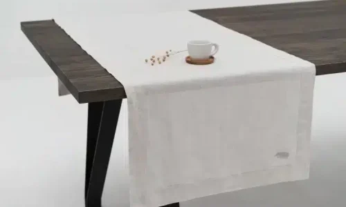 A dark wood table with black legs is covered with a white linen table runner from renowned linen weavers. On the runner are a white cup and saucer as well as a small dried plant. The background is white and minimalist.