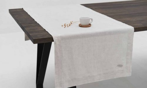A dark wooden table with the Natural linen tablecloth by Geniksa as a light beige runner, paired with a white cup and saucer and a small dried plant. The minimalist white background underscores this elegant ambience.