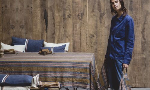Eine Person in einer blauen Jacke steht an einem Tisch, der mit Olympia Stripe - reines Leinen für den Tisch von Libeco gedeckt ist, und hält ein passendes Tuch. Holzwände und Kissen auf einer nahen Bank vervollständigen die Szene.