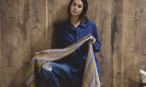 Eine Frau in blauem Jeanshemd und Jeans steht an einer Holzwand, hält das Olympia - reines Leinenhandtuch von Libeco in der Hand und blickt nachdenklich zur Seite.