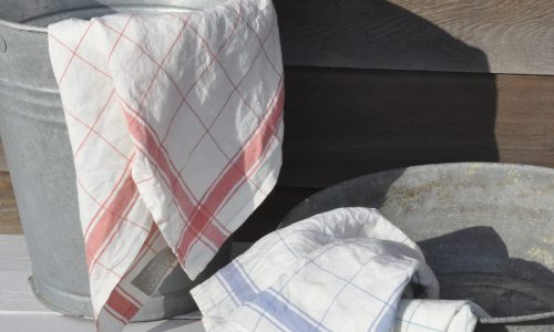 Zwei Metallwaschwannen stehen im Freien; in der einen ist ein weißes Tuch mit roten Streifen, in der anderen ein weißes Tuch mit blauen Streifen drapiert.