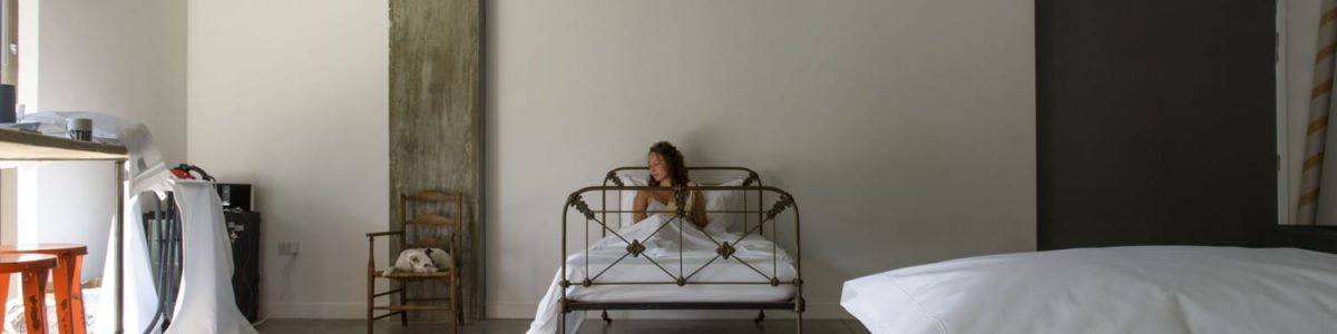 Eine Frau sitzt auf einem Metallbett, eingewickelt in weiße Laken, in einem minimalistischen Schlafzimmer mit einer Betonsäule, einem Holzstuhl und verstreuten Möbeln. Der Raum ist geräumig und lichtdurchflutet.