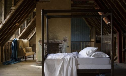 Das rustikale Dachgeschoss-Schlafzimmer verfügt über ein hölzernes Himmelbett, das mit Prager Percal 400TC Bettwäsche von Peter Reed bezogen ist. Ein blauer Sessel neben freiliegenden Balken und einem abblätternden Kamin vervollständigen den Vintage-Charme des Zimmers.