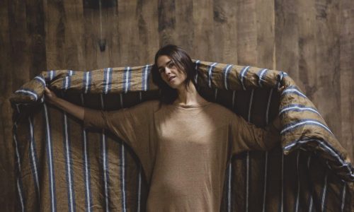 Eine Frau in einem weiten braunen Kleid steht an einer Holzwand und hält Salem Leinen Bettwäsche von Libeco, ein großes, gestreiftes Leinenbettwäsche-Set, mit ausgestreckten Armen hinter sich.