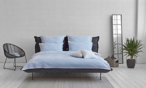 Ein modernes Schlafzimmer mit einem hellblauen Bett, einem schwarzen Kopfteil, zwei Kissen, einem schwarzen Drahtstuhl, einem hohen Spiegel, einer Topfpflanze und einem kleinen grauen Teppich auf weißem Holzboden vor einer weißen Backsteinwand.