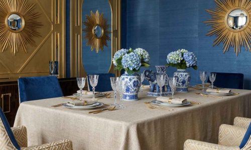 Elegantes Esszimmer mit Salome Reinleinen-Tischdecken von Schlitzer Leinen, blauem und goldenem Dekor, blauen Hortensien in gemusterten Vasen, goldenen Wandspiegeln mit Sonnenschliff, blauen Wänden, gemusterten Stühlen und einem auffälligen Kristalllüster.